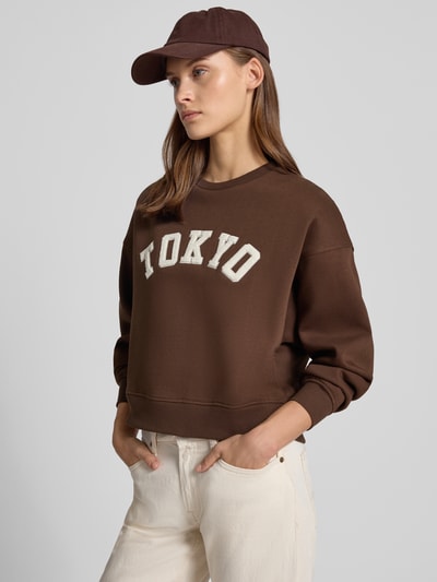 Vrouwenmodel draagt relaxed fit dames sweatshirt van katoenmix Model 'Tatum' van JJXX, statementpatch, ribboordjes, met lange mouwen, ronde hals, detailopname bovenlichaam.