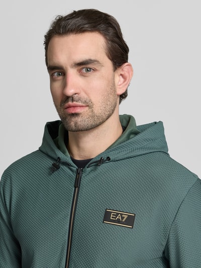 Männermodel trägt Regular Fit Sweatjacke mit Logo-Patch von EA7 Emporio Armani, grün, Strukturmuster, Nahaufnahme Oberkörper.