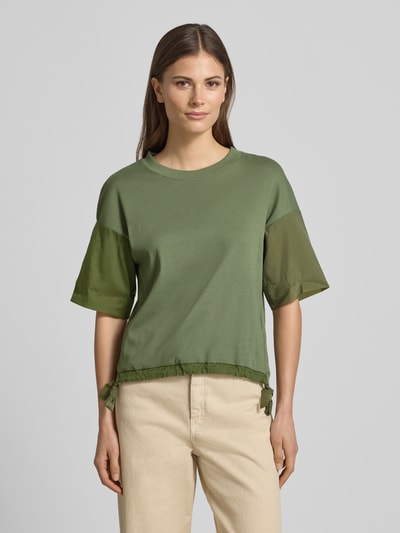 Kobieta nosi t-shirt z okrągłym dekoltem i tunelem od Marc Cain, mieszanka bawełny i elastanu, krój relaxed fit, zakładki na rękawach, długość tyłu 51 cm w rozmiarze 36, damski t-shirt; widok całej sylwetki z przodu.