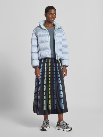 Frauenmodel trägt Regular Fit Jacke in Stepp-Optik mit Stehkragen von Marc Cain, hellblau, kombiniert mit plissiertem Rock, elastische Abschlüsse, Zweiwege-Reißverschluss, Produktdetails: Rückenlänge 50 cm, Schulterbreite 39 cm, Brustweite 57 cm, Ärmellänge 52 cm, Ganzkörperansicht von vorne.