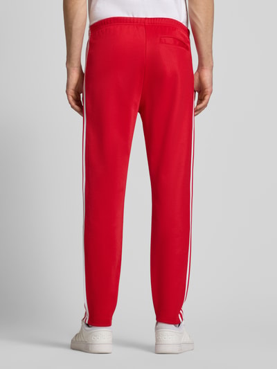 Männermodel trägt Sweatpants mit elastischem Bund von adidas Originals in Rot mit weißen Streifen, Baumwoll-Mix, lockerer Schnitt, Gesäßtasche, Ganzkörperansicht Rückansicht.