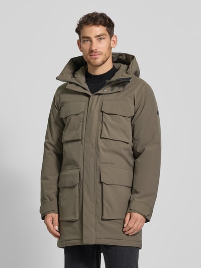 Męski model nosi Parka mit Pattentaschen Modell 'AIDEN' od Didriksons, kolor oliwkowy, regular fit, kaptur, zdjęcie całej sylwetki z przodu. Długość 92 cm, szerokość ramion 47 cm, szerokość klatki 56 cm, długość rękawa 69 cm.