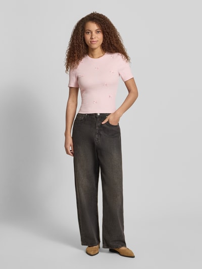 Modelka nosi różowe Slim Fit T-Shirt aus Baumwoll-Mix Modell 'FEMKE' od Only, jednolity kolor, drobny prążek, ozdobne kokardki, krótki rękaw, okrągły dekolt, widok całej sylwetki z przodu.