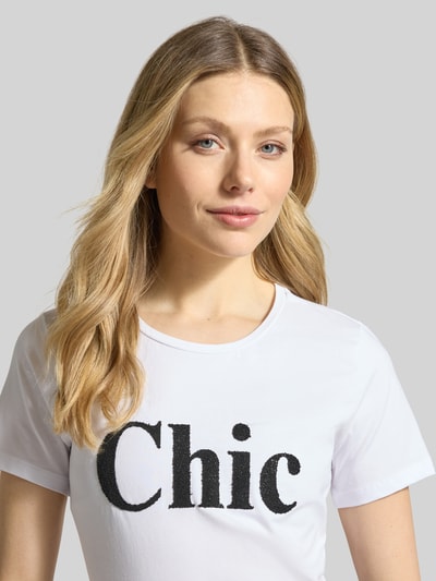 Frauenmodel trägt T-Shirt mit Rundhalsausschnitt und Perlen von More & More, weiß, Statement-Print, Baumwoll-Elasthan-Mix, Nahaufnahme Oberkörper.