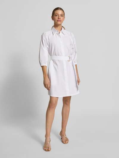Frauenmodel trägt ausgestelltes, weißes Hemdblusenkleid aus reiner Baumwolle Modell 'KILONI' von HUGO mit Bindegürtel, Ganzkörperansicht von vorne.