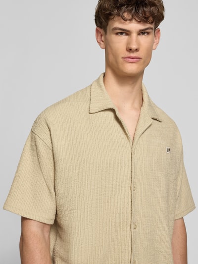 Männermodel trägt Regular Fit Freizeithemd mit Label-Patch Modell 'LIBCO' von Pegador, beigefarbener Material-Mix, Knopfleiste, Reverskragen, Kurzärmelig, Struktur-Oberfläche, Nahaufnahme Oberkörper. Rückenlänge bei Größe M: 72 cm, Schulterbreite: 41 cm, Ärmellänge: 35 cm.