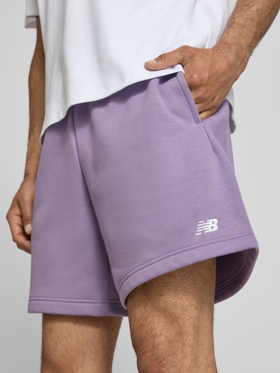 Männermodel trägt Relaxed Fit Shorts mit Logo-Stitching von New Balance aus Baumwoll-Mix in Flieder, sichtbarer Tunnelzugbund, Nahaufnahme Oberschenkel.