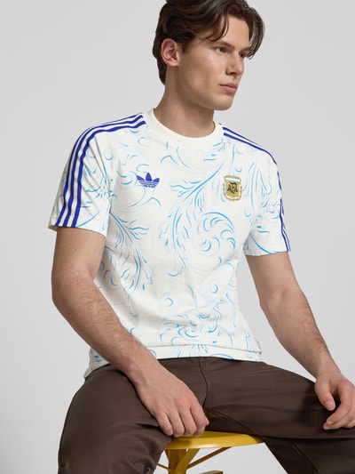 Männermodel trägt Regular Fit Trikot aus reiner Baumwolle von Adidas Originals mit Optik:Allover-Muster, Logo-Stitching, Kontraststreifen, Rundhals, Kurzarm, Materialverarbeitung: Jersey, Ganzkörperansicht von vorne.