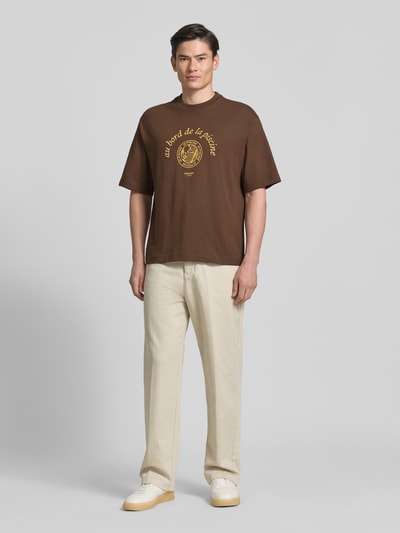 Männermodel trägt Relaxed Fit T-Shirt aus reiner Baumwolle Modell 'BOXYNORMAN GRAPHIC' von SELECTED HOMME in Braun mit Motiv-Print, Rundhalsausschnitt und kurzen Ärmeln, Ganzkörperansicht von vorne.