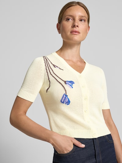 Frauenmodel trägt Slim Fit Sweatjacke aus Baumwoll-Mix Modell 'RANCH' von MAX&Co. in Strickoptik, gelb mit blauen floralen Intarsien, einreihige Knopfleiste, Halbarm, V-Ausschnitt, Nahaufnahme Oberkörper.