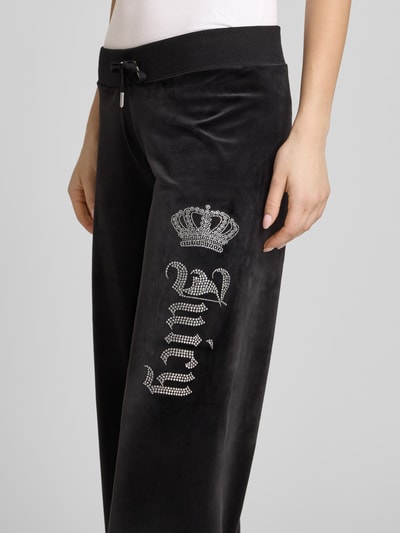 Kobiece modelka nosi Sweatpants mit Ziersteinbesatz Modell 'GOTHIC CROWN' von Juicy Couture, czarny, prosty krój, materiał z domieszką stretchu, elastyczny pas z troczkiem, zdobienia z kamieni, damskie spodnie dresowe, zbliżenie na biodra i uda, kontekst: długość wewnętrzna nogawki w rozmiarze S: 78 cm.