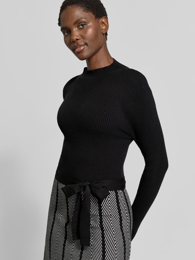 Kobiece modelka nosi sukienkę 'Knielanges Kleid im 2-in-1-Look' od Apricit, mieszanka wiskozy, krój o linii a, długość do kolan, efekt 2 w 1, wiązany pasek, długość tyłu w rozmiarze S: 105 cm, szerokość w rozmiarze S: 39 cm, długość rękawów w rozmiarze S: 64 cm, czarna góra, wzorzysta spódnica, faktura prążkowana, geometryczne wzory, zdjęcie półzbliżenie od przodu.