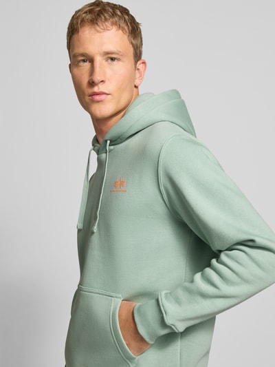 Męski model nosi Hoodie mit Logo und Känguru-Tasche von Alpha Industries w kolorze miętowym, mieszanka bawełny, krój regular fit, długość tyłu 69 cm, długość rękawów 64 cm, logo, kieszeń kangurka, zbliżenie na górną część ciała z przodu.
