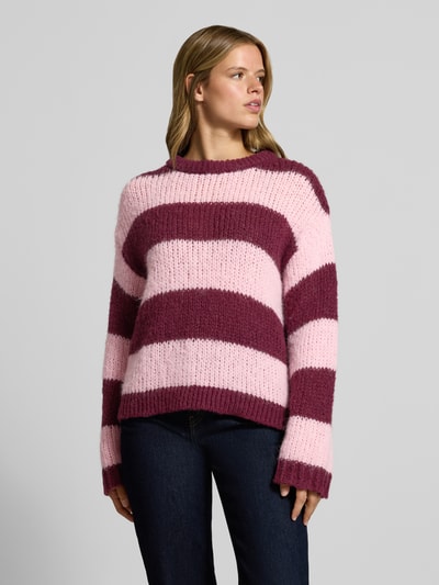 Frauenmodel trägt Relaxed Fit Pullover im Chunky Knit Look Modell 'DORTHEA' von PIECES, breite rosa und bordeauxrote Streifen, grobe Stricktextur, Ganzkörperansicht von vorne. Damen Pullover von PIECES.
