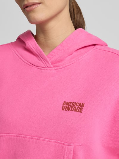 Kobiece model prezentuje bluzę z kapturem z logo Hoodie mit Känguru-Tasche Modell 'PLIZZY' od American Vintage z bawełny, krój relaxed fit, kieszeń kangurka, kolor różowy, logo, zbliżenie na górną część torsu