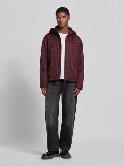 Männermodel trägt Regular Fit Jacke mit Allover-Logo-Print von Emporio Armani in Dunkelrot, dezenter Glanz, Zweiwege-Reißverschluss, Ganzkörperansicht von vorne. Kontext: Damen Jacke von Armani, Logo-Applikation, Kapuze mit Tunnelzug, Eingrifftaschen, Rückenlänge 61 cm, Schulterbreite 47 cm, Brustweite 58 cm, Ärmellänge 65 cm.