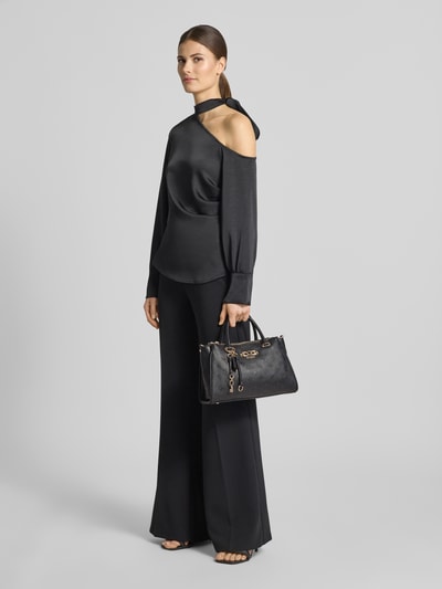 Frauenmodel trägt Handtasche mit Allover-Logo-Prägung Modell 'Anise' von Guess, in Schwarz, mit Schulterriemen, Magnetverschluss, Ganzkörperansicht von vorne.
