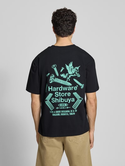 Mężczyznamodel nosi męski t-shirt z nadrukiem i okrągłym dekoltem od Edwin, czarna bawełna, krój relaxed fit, długość tyłu 72 cm, nadruk w zielonym kolorze, widoczne elementy graficzne, tekstura gładka, całkowity widok tyłem.