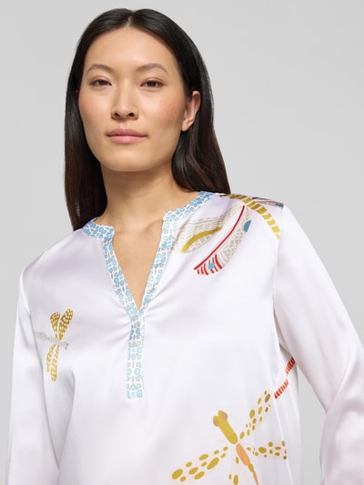 Kobiece modelka ma na sobie Relaxed Fit Tunika-Bluse mit Allover-Motiv-Print od HERZENS, dekolt z lamówką, krój relaxed fit, nadruk z motywem na całej powierzchni, z długim rękawem, tył dłuższy; kolor biały z kolorowymi wzorami, gładka tekstura. Zbliżenie na górną część ciała z przodu.