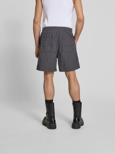 Männermodel trägt Shorts mit Stretch-Anteil von Forét in Grau mit feinem Streifenmuster, aufgesetzten Gesäßtaschen, Ganzkörperansicht Rückansicht.