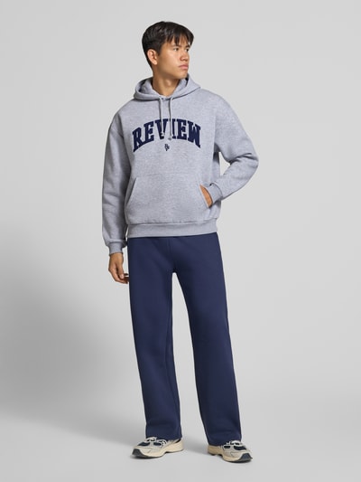 Männermodel trägt Hoodie mit Label-Stitching von REVIEW in Grau und Dunkelblau, Baumwollmischung, Känguru-Tasche, Ganzkörperansicht von vorne.