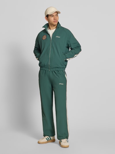 Männermodel trägt Trackjacket mit Label-Stitching Modell 'Slipstream' von PROHIBITED in Dunkelgrün mit Kontraststreifen, Ganzkörperansicht von vorne. Relaxed Fit, Stehkragen, Reißverschluss, Rückenlänge bei Größe M 64 cm, Ärmellänge 73 cm.