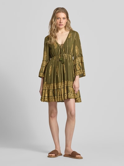 Frauenmodel trägt Minikleid aus Viskose-Mix mit Schnürung von Apricot in olivgrün mit goldfarbenem Muster, A-Linie, V-Ausschnitt, Trompetenärmel, Ganzkörperansicht von vorne, Rückenlänge 83 cm, Brustweite 35 cm, Ärmellänge 50 cm.