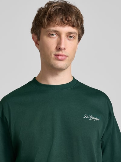 Männermodel trägt Loose Fit T-Shirt aus reiner Baumwolle Modell 'LES CLASSIQUES' von Only & Sons, Optik: Unifarben in Dunkelgrün, mit Schriftzug, Rundhals, Kurzarm, Jersey, Nahaufnahme Oberkörper.