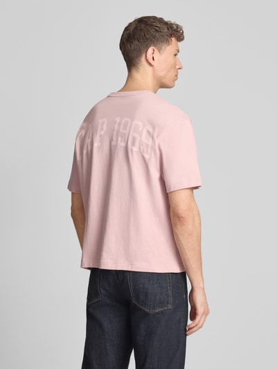 Męski model nosi T-Shirt mit Logo und Rundhalsausschnitt od GAP, bawełna, krój regular fit, logo, okrągły dekolt, długość tyłu w rozmiarze M: 61 cm, kolor jasnoróżowy, proste kształty, jednolita tekstura, całkowity widok z tyłu.