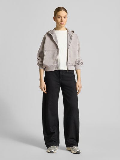 Frauenmodel trägt Sweatjacke mit Zweiwege-Reißverschluss Modell 'RANERA' von Drykorn aus Reine Baumwolle im Relaxed-Fit mit Kapuze, gedeckte Farben, kastige Form, samtige Textur, Ganzkörperansicht von vorne.