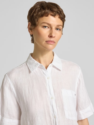 Kobieta modelka nosi Loose Fit Leinenbluse mit Brusttasche od Tommy Hilfiger, biała, lniana tekstura, kieszeń na piersi, krój loose fit, haftowane logo, kołnierzyk koszulowy, krótki rękaw, zbliżenie na górną część ciała