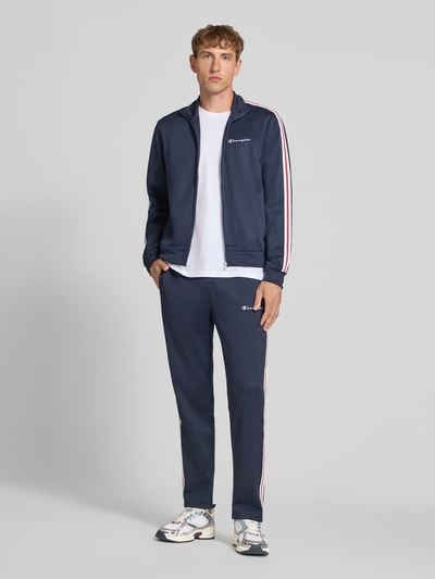 Männermodel trägt dunkelblaue Sweatpants mit Bordüre von Champion, gerader Schnitt, mit Seitentaschen, weiße Streifen, Ganzkörperansicht von vorne.