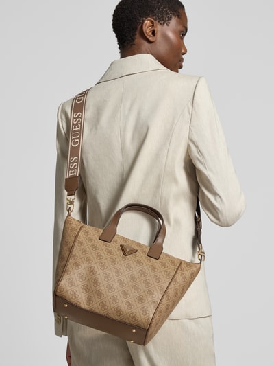 Männermodel trägt Tote Bag mit Label-Patch Modell 'FOLLIE' von Guess, beżowy z brązowymi uchwytami, regulowany pasek na ramię, logo marki, główna przegroda z zamkiem błyskawicznym, kieszeń zapinana na zamek błyskawiczny wewnątrz, szerokość w rozmiarze 1: 36 cm, długość w rozmiarze 1: 24 cm, głębokość w rozmiarze 1: 12 cm, długość paska na ramię w rozmiarze 1: 114 cm, Ganzkörperansicht Rückansicht.