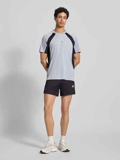 Męski model trägt T-shirt mit Label-Print von adidas Training, jasno-szary z czarnymi detalami, krój regular fit, okrągły dekolt, nadruk z logo, długość tyłu 72 cm w rozmiarze M, Ganzkörperansicht von vorne.