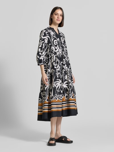Frauenmodel trägt Midikleid mit Eingrifftaschen und 3/4-Arm von Emily Van den Bergh, Reine Baumwolle, A-Linie, Allover-Look, V-Ausschnitt, Knopfleiste, schwarze und weiße Muster, orangefarbene und beigefarbene Blockstreifen am Saum, fließender Stoff, Ganzkörperansicht von vorne. Rückenlänge Größe 36: 126 cm, Brustweite: 43 cm, Ärmellänge: 46 cm.