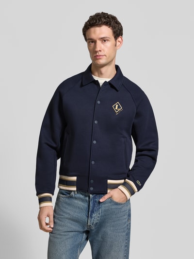 Männermodel trägt Regular Fit Sweatjacke aus Baumwoll-Mix von Lacoste in Dunkelblau mit goldenen Details, Ganzkörperansicht von vorne. Herren Sweatjacke von Lacoste