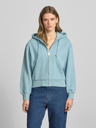 Modelka kobieca nosi błękitną bluzę rozpinaną z bawełny, krój loose fit, model 'Casey' od Carhartt Work In Progress z wyhaftowanym logo, kapturem, zamkiem błyskawicznym, wpuszczanymi kieszeniami i elastycznymi wykończeniami, widok całej sylwetki z przodu.
