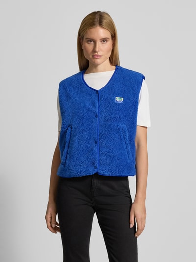 Vrouwenmodel draagt felblauwe Bodywarmer met ronde hals Model 'HOKTOWN' van American Vintage, teddybont, drukknoopsluiting, steekzakken, labeldetail, los model, voorkant bovenlichaam close-up.