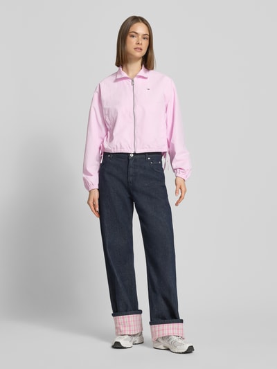 Frauenmodel trägt Relaxed Fit Blouson mit Taillenbändern von Tommy Jeans in Hellrosa, unifarben, Stehkragen, Reißverschluss, Logo-Stitching, seitliche Eingrifftaschen, elastische Ärmelabschlüsse, kombiniert mit dunkelblauer Hose und Sneakers, Ganzkörperansicht von vorne.