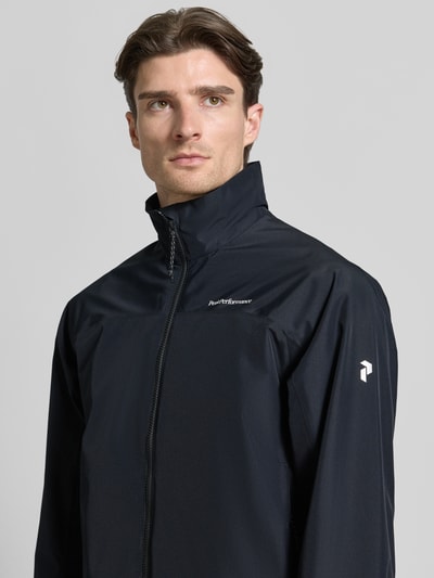 Męski model nosi Regular Fit Jacke mit Logo-Print Modell 'Quest' od PEAK PERFORMANCE, kolor czarny, stójka, zamek, zbliżenie na górną część ciała. Długość pleców w rozmiarze M: 74 cm, szerokość ramion w rozmiarze M: 45 cm, szerokość w rozmiarze M: 55 cm, długość rękawa w rozmiarze M: 65 cm.