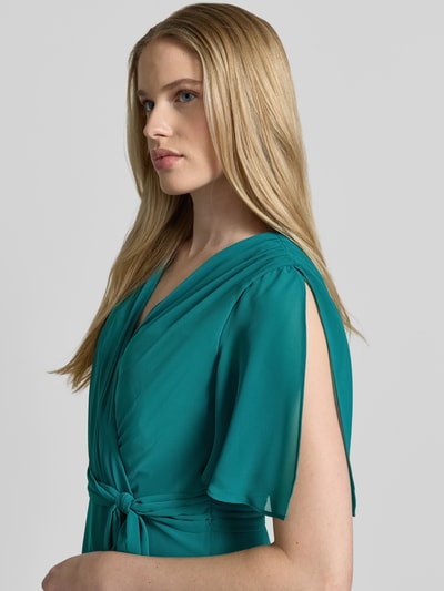 Frauenmodel trägt Cocktailkleid in Wickel-Optik mit Volantsaum von Christian Berg Cocktail in Türkis, Chiffon, Schmetterlingsärmel, Bindegürtel, Midikleid, Nahaufnahme Oberkörper.