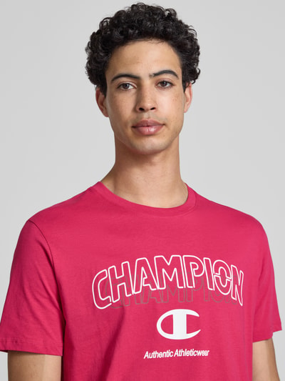 Männermodel trägt T-Shirt mit Label-Print und Rundhalsausschnitt von CHAMPION in Rot, reine Baumwolle, Regular Fit, Nahaufnahme Oberkörper.