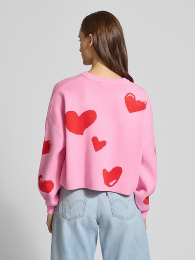 Frauenmodel trägt Oversized Strickpullover aus Viskose-Mix Modell 'SHYLA' von Noisy May in Pink mit roten Herz-Motiven, Ballonärmeln, elastische Ärmelabschlüsse, Statement-Schriftzug, Rückenlänge 44 cm, Ganzkörperansicht Rückansicht.
