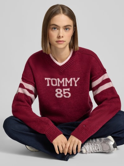 Kobiece modelka nosi Oversized Strickpullover mit Woll-Anteil od Tommy Jeans w kolorze bordowym z białymi napisami i paskami, dzianina, swobodny fason, dekolt w serek, długie rękawy, widoczny label, zdjęcie w zbliżeniu na górną część ciała.
