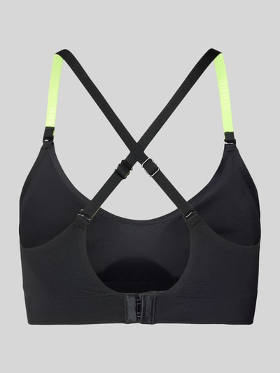 Frauenmodel trägt Triangel-BH mit Logo und gekreuzten Trägern von PUMA, Schwarz mit neongelben Verstellbarer-Trägern, Material-Mix mit Stretch-Anteil, Hakenverschluss, Körpernahe Passform, Ganzkörperansicht Rückansicht.