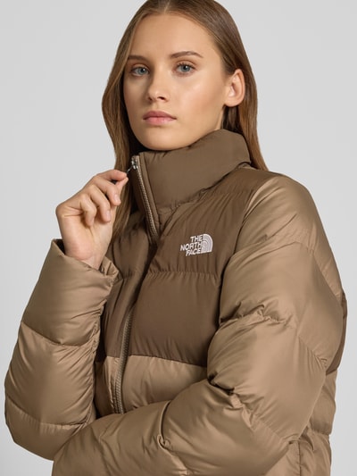 Kobiece modelka nosi kurtkę pikowaną Model 'SAIKURU' od The North Face, brązowe odcienie, stójka, wyhaftowane logo, zamek błyskawiczny, kieszenie zapinane na zamek błyskawiczny, krój regular fit, detaliczna tekstura, Szerokość w rozmiarze S: 53 cm, Szerokość ramion w rozmiarze S: 43 cm, Długość tyłu w rozmiarze S: 59 cm, Długość rękawów w rozmiarze S: 62 cm, zbliżenie na górną część ciała