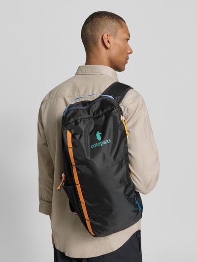 Männermodel trägt Rucksack mit Logo-Print Modell 'Batac' von Cotopaxi in Schwarz mit bunten Details, Hauptfach mit Reißverschluss, Rückenansicht. Breite 22 cm, Länge 48 cm, Tiefe 15 cm, Länge Schulterriemen 65 cm, verstellbare Trageriemen, Herren Rucksack.