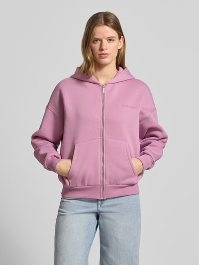 Frauenmodel trägt Sweatjacke mit Logo und Känguru-Tasche von Review in Rosa aus Baumwoll-Mix, Relaxed-Fit, mit Kapuze, Reißverschluss und Känguru-Tasche. Ganzkörperansicht von vorne.