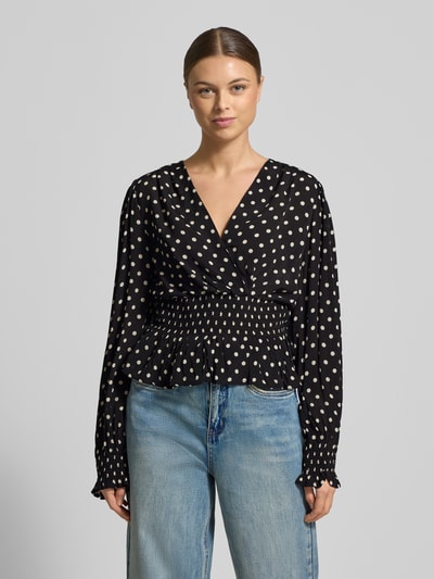 Frauenmodel trägt Bluse aus reiner Viskose in Wickel-Optik Modell 'Tamona' von Neo Noir, schwarz mit weißen Punkten, V-Ausschnitt, Regular Fit, taillierte Passform, Rüschenbesatz, langärmeliges Design, strukturierte Textur, Ganzkörperansicht von vorne.