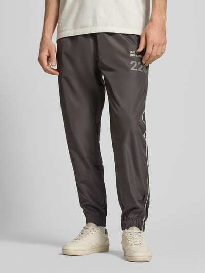 Männermodel trägt Trainingshose mit elastischem Bund von HALO in Grau mit Label-Print, Relaxed Fit und Reißverschlusstaschen, Nahaufnahme Unterkörper. Innenbeinlänge bei Größe M: 40 cm.